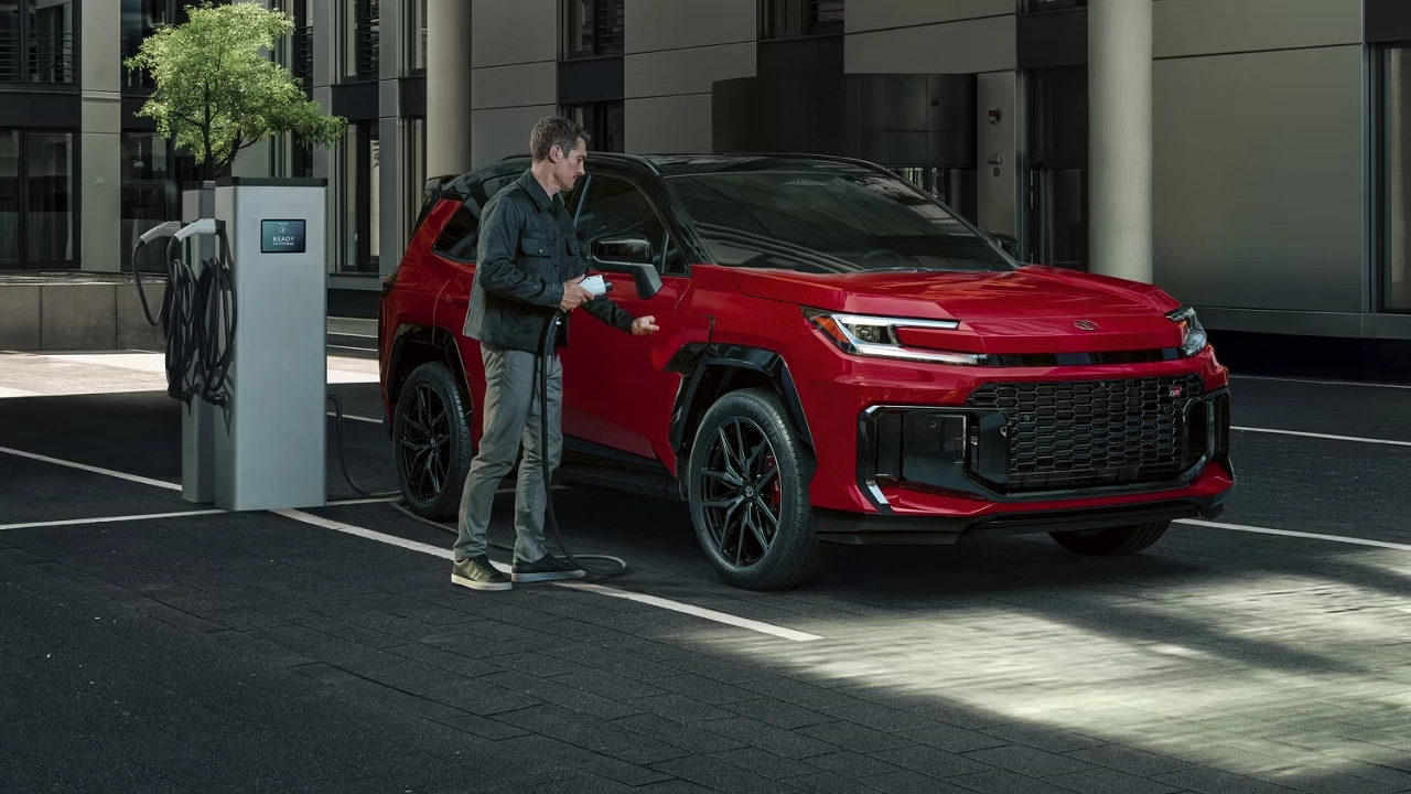 A man plugs a charging cable into a red Toyota RAV4 at a public charging station, demonstrating the SUV's range.