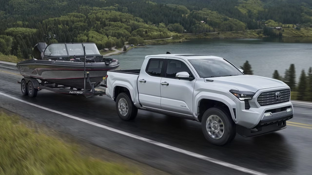 A white 2026 Toyota Tacoma towing a boat on a road along a lake