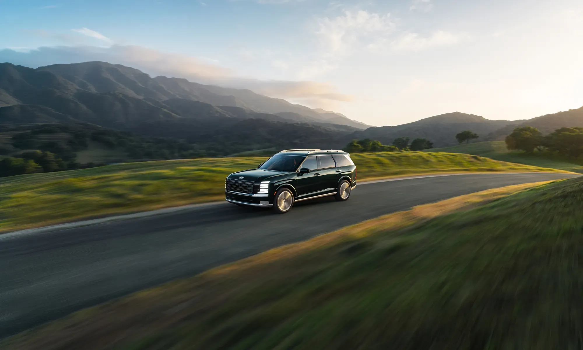 Green 2027 Hyundai Palisade Hybrid driving on a road at the foot of the mountains.