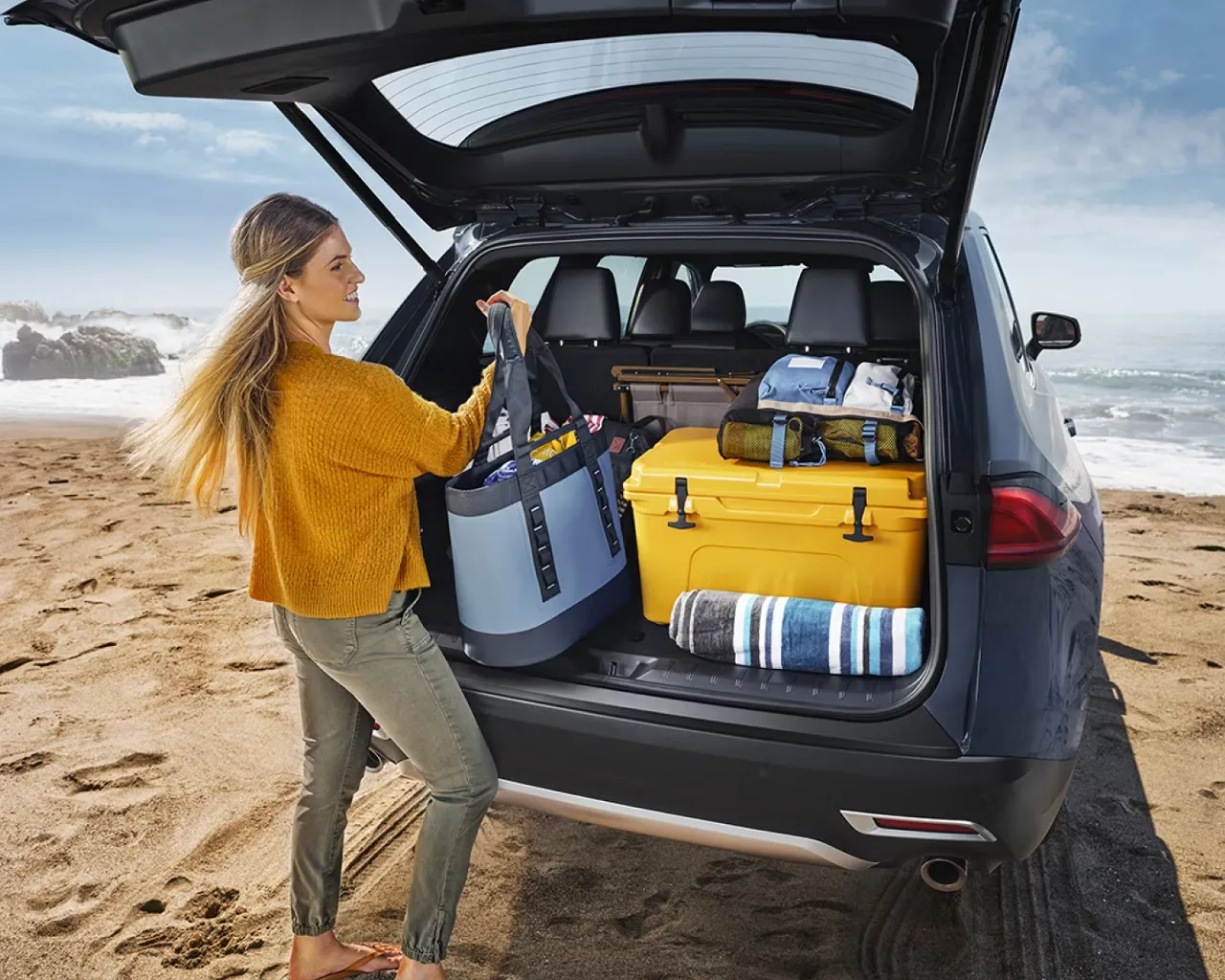 Woman loading luggage into the open cargo area of a 2026 Toyota Grand Highlander Hybrid at the beach.